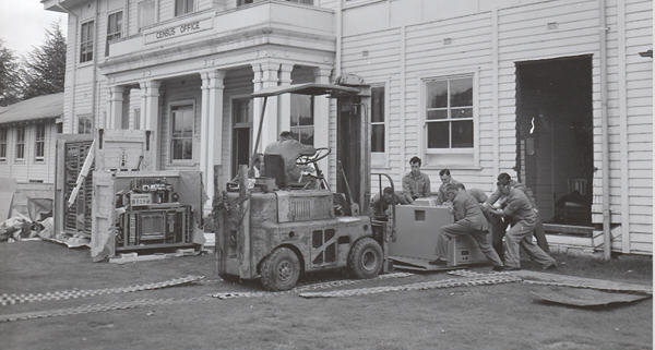 Six men in overalls assist a forklift driver in loading a Census Trio machine  so that he canl lift it through a large hole cut in the building