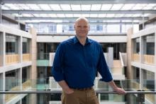 Mark Harding leans one arm on a railing on a platform overlooking a large office building. He is smiling directly at the camera
