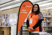 a census staff member wearing an orange shirt smiles at the camera in a community library. a banner which reads 'census here to help' can be seen in the background
