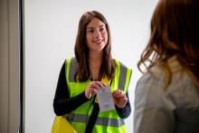 a census staff member wearing a yellow high-vis vest greets a woman at the door of an apartment. she is displaying a Census ID badge