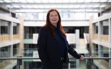 Jenny Telford leans one arm on a railing on a platform overlooking a large office building. She is smiling directly at the camera